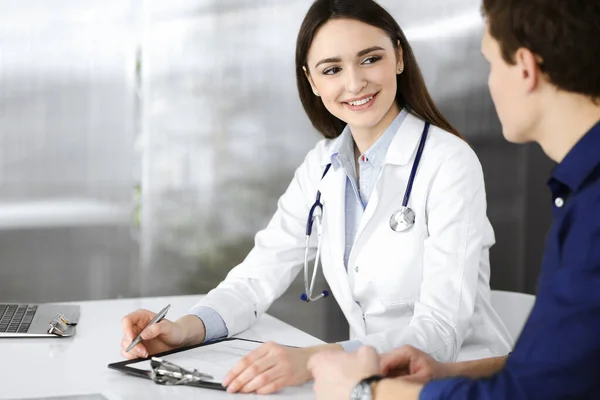 Freundliche junge Ärztin hört ihrer Patientin zu, während sie zusammen am Schreibtisch in einem Schrank sitzen. Ärztin hält Klemmbrett zum Auffüllen von Medikamenten in der Hand — Stockfoto