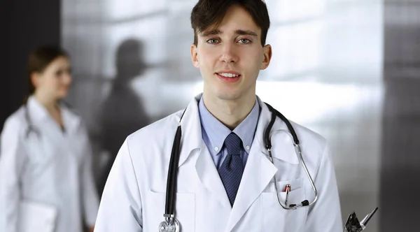 현대 의학에서 클립보드를 들고 서 있는 친절 한 젊은 남성 의사. 여성 동료는 의사의 배경으로 약학 기록을 작성하느라 바쁩니다. 의학적 개념 — 스톡 사진
