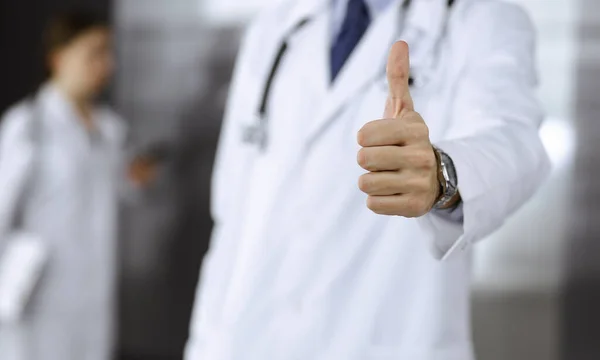 Onbekende man-arts die rechtop staat en OK teken toont met duim omhoog in de moderne kliniek. Vrouwelijke collega heeft haast op de achtergrond van de arts. Geneesmiddelenconcept — Stockfoto