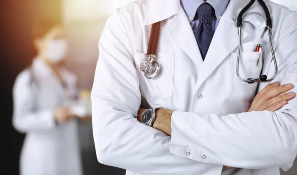 Güneşli klinikte kolları çapraz duran koruyucu maskeli kimliği belirsiz erkek doktor. Coronavirüs salgını sırasında ilaç kavramı
