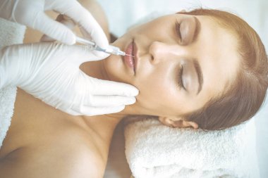 Güzellik uzmanı güneşli klinikte genç esmer kadının yüzüne şırıngayla güzellik prosedürü uyguluyor. Kozmetik ilaç ve ameliyat, güzellik iğneleri.