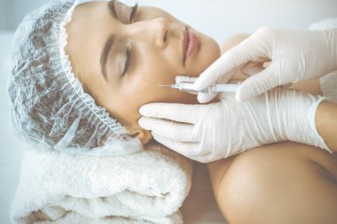 Güzellik uzmanı güneşli klinikte genç esmer kadının yüzüne şırıngayla güzellik prosedürü uyguluyor. Kozmetik ilaç ve ameliyat, güzellik iğneleri.