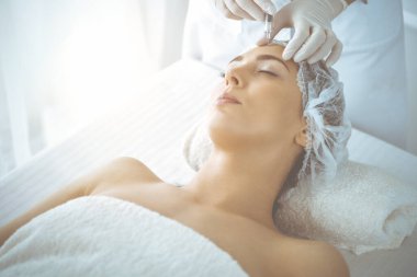 Güzellik uzmanı güneşli klinikte genç esmer kadının yüzüne şırıngayla güzellik prosedürü uyguluyor. Kozmetik ilaç ve ameliyat, güzellik iğneleri.