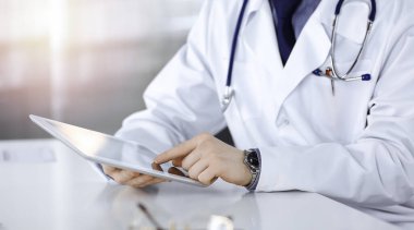 Bilinmeyen erkek doktor, tablet bilgisayarla oturuyor ve çalışıyor. Karanlık bir klinikte, arka planda ışık parıltısı, ellerin yakın çekimi.