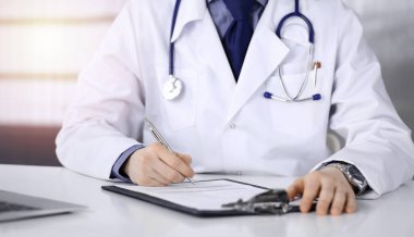 Bilinmeyen bir erkek doktor, karanlık bir klinikte tıbbi geçmişi olan bir panoyla oturuyor ve çalışıyor. Arka planda ışık parıltısı var.