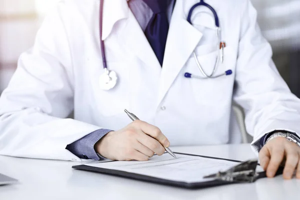 Médico masculino desconocido sentado y trabajando con portapapeles del historial de medicamentos en una clínica oscura, resplandor de luz en el fondo, primer plano de las manos — Foto de Stock