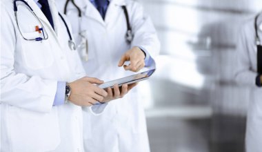 Bilinmeyen bir grup doktor hastane ofisinde dururken bazı tıbbi isim kayıtlarını kontrol etmek için bir bilgisayar tableti kullanıyor. Doktorlar hastaları muayene etmeye ve yardım etmeye hazır. Tıbbi yardım, sigorta