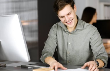 Yeşil gömlekli genç işadamı ve programcı arka planda bir bayan meslektaşıyla birlikte modern bir dolapta otururken defterini çeviriyor ve kameraya bakıyor.