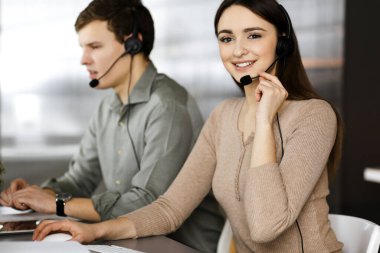 Kulaklık takmış iki genç bir ofiste masada otururken müşterilerle konuşuyor. Kadınlara odaklan. İş yerindeki çağrı merkezi operatörleri