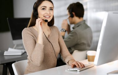 Modern bir ofiste meslektaşıyla birlikte masa başında otururken, kulaklıklı genç bir kız bir firma müşterisiyle konuşuyor. İş yerindeki çağrı merkezi operatörleri