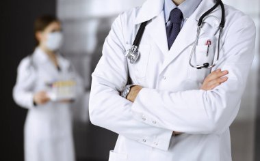 Koruyucu maskeli kimliği belirsiz erkek doktor klinikte kollarını kavuşturmuş duruyor. Coronavirus salgını sırasında ilaç konsepti. Covid 2019