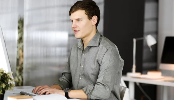 Un jeune homme d'affaires prospère et programmeur en chemise verte travaille dur sur son ordinateur, tout en étant assis sur le bureau dans un cabinet moderne. Prise de vue ou portrait d'entreprise dans un bureau — Photo