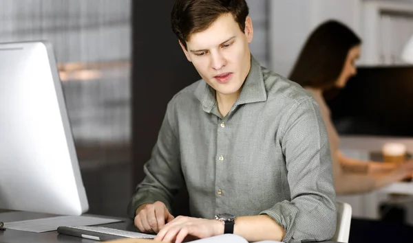 Young businessman and programmer in a green shirt is checking a business plan, while sitting at the desk in a modern cabinet together with his female colleague on the background. Concept of successful