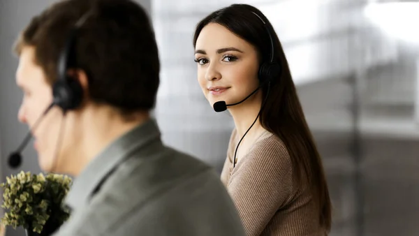 Joven chica alegre en auriculares está mirando a la cámara, mientras está sentado en el escritorio con un colega masculino. Retrato del operador del centro de llamadas en el trabajo —  Fotos de Stock