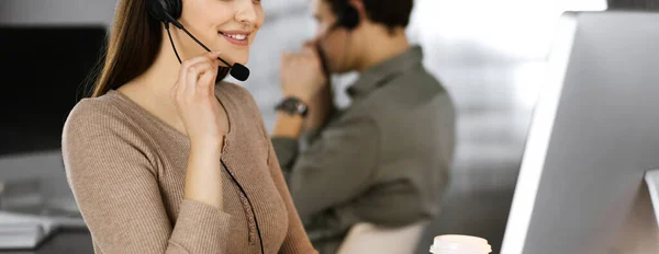 Jeune fille amicale dans les écouteurs parle à un client des entreprises, tout en étant assis sur le bureau dans un bureau moderne avec son collègue. Opérateurs de centre d'appels au travail — Photo