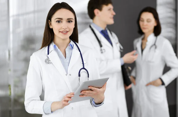 Sorridente intelligente donna-medico sta tenendo in mano un tablet, mentre lei è in piedi insieme ai suoi colleghi in una clinica. Medici al lavoro. Servizio medico perfetto in ospedale — Foto Stock