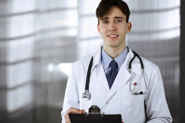 Vriendelijke jonge mannelijke arts staat met klembord in moderne kliniek. Geneesmiddelenconcept — Stockfoto