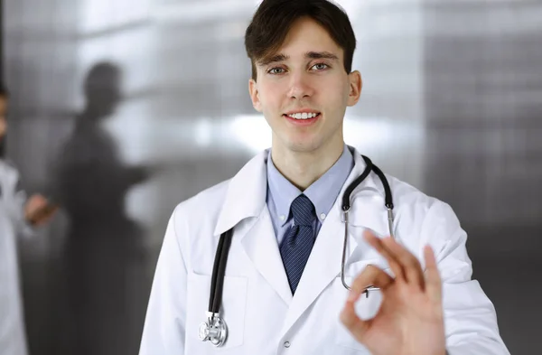 Freundlicher Mann-Arzt, der gerade steht und in der Klinik Ok-Zeichen zeigt. Die Kollegin hat es eilig im Hintergrund des Arztes. Medizinkonzept — Stockfoto