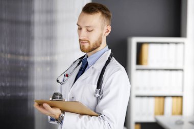 Kızıl sakallı doktor çalışma yerinin yanındaki klinikte dikiliyor. Hekim portresi. Tıp ve sağlık hizmetleri kavramı