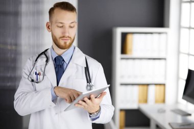 Dost canlısı kızıl sakallı doktor çalıştığı yerdeki klinikte tablet bilgisayar kullanıyor. Hastane ve tıp konseptinde mükemmel bir tıbbi hizmet.