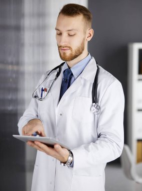 Dost canlısı kızıl sakallı doktor çalıştığı yerdeki klinikte tablet bilgisayar kullanıyor. Hastane ve tıp konseptinde mükemmel bir tıbbi hizmet.