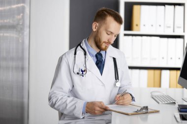 Dost canlısı kızıl sakallı doktor klinikteki panoda oturuyor ve yazıyor. Hastanede mükemmel bir tıbbi hizmet. Tıp konsepti