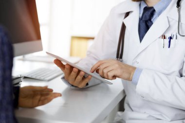 Bilinmeyen kızıl sakallı doktor ve hasta kadın güncel sağlık muayenesini güneşli klinikte otururken ve tablet bilgisayar kullanırken tartışıyorlar, yakın plan. Tıp konsepti