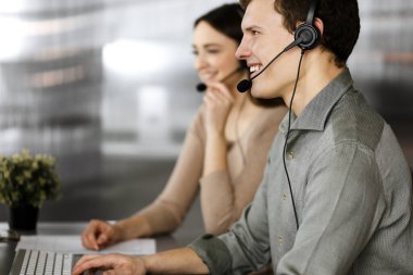 Modern bir ofiste masada yan yana otururken, kulaklıklı iki genç müşterilerle konuşuyor. İnsanoğluna odaklan. İş yerindeki çağrı merkezi operatörleri