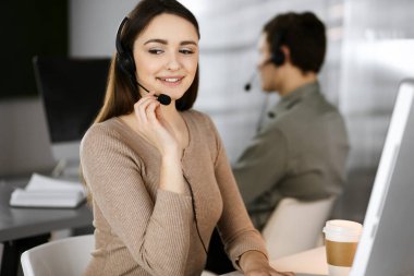 Modern bir ofiste meslektaşıyla birlikte masa başında otururken, kulaklıklı genç bir kız bir firma müşterisiyle konuşuyor. İş yerindeki çağrı merkezi operatörleri