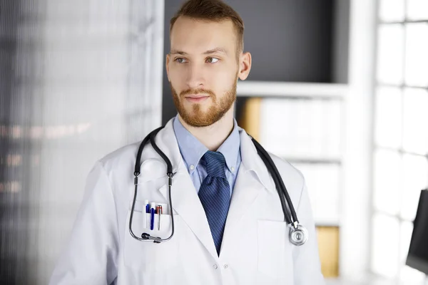 Medico con la barba rossa in piedi dritto in clinica vicino al suo posto di lavoro. Ritratto di medico. Medicina e concetto di assistenza sanitaria — Foto Stock