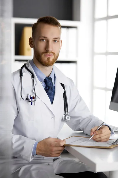 Amigável médico de barba vermelha sentado e escrevendo na área de transferência na clínica. Serviço médico perfeito no hospital. Conceito de medicina — Fotografia de Stock