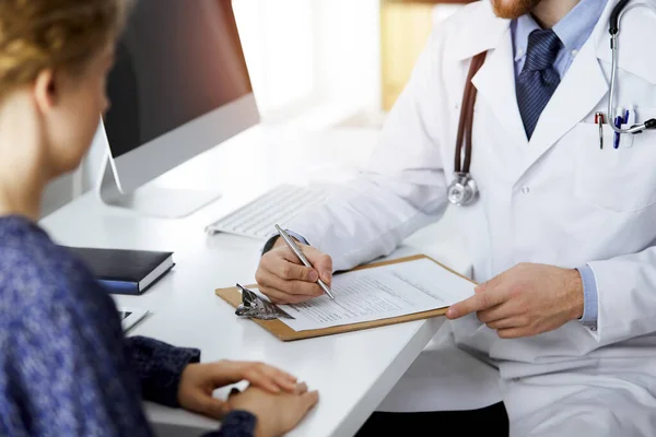 Amistoso médico de barba roja y mujer paciente discutiendo el examen de salud actual mientras está sentado en una clínica soleada. Concepto de medicina — Foto de Stock