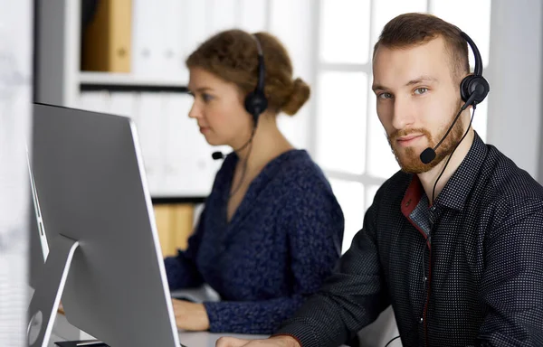 Homme d'affaires à barbe rouge parlant par casque près de sa collègue féminine alors qu'il était assis dans un bureau moderne. Groupe de personnes diversifiées au centre d'appels. Télémarketing et service à la clientèle — Photo