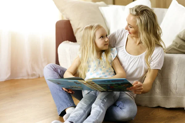 Glückliche Familie. Blonde junge Mutter liest ihrer süßen Tochter ein Buch vor, während sie am Holzboden in einem sonnigen Raum sitzt. Mutterschaftskonzept — Stockfoto