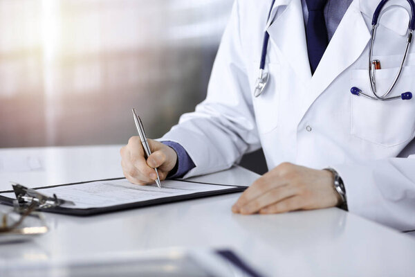 Unknown male doctor sitting and working with clipboard of medication history record in a darkened clinic, glare of light on the background, close-up of hands