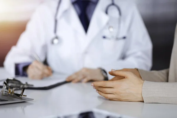 Médico desconhecido e mulher paciente discutindo algo enquanto se senta em uma clínica escurecida, brilho de luz no fundo. Close-up de mãos — Fotografia de Stock