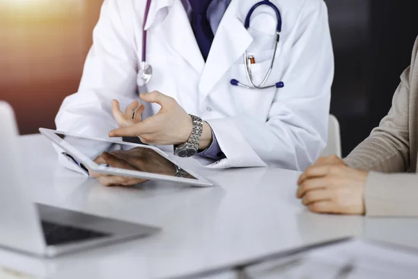 Unknown male doctor and patient woman discussing something while using tablet computer in a darkened clinic, glare of light on the background. Best medical service in hospital, medicine, pandemic stop