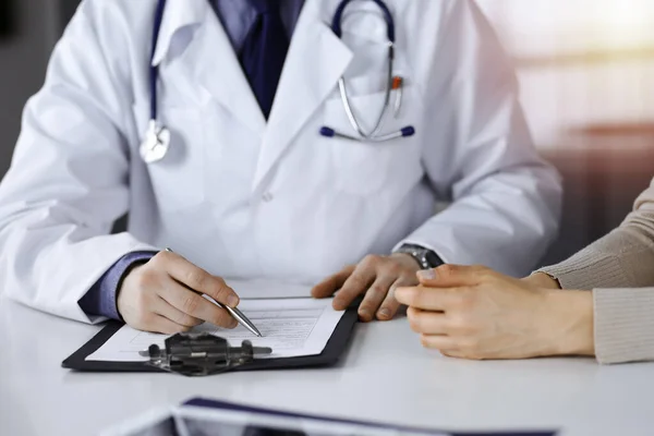 Médico masculino desconocido y mujer paciente discutiendo algo mientras está sentado en una clínica oscura y usando portapapeles, resplandor de luz en el fondo — Foto de Stock