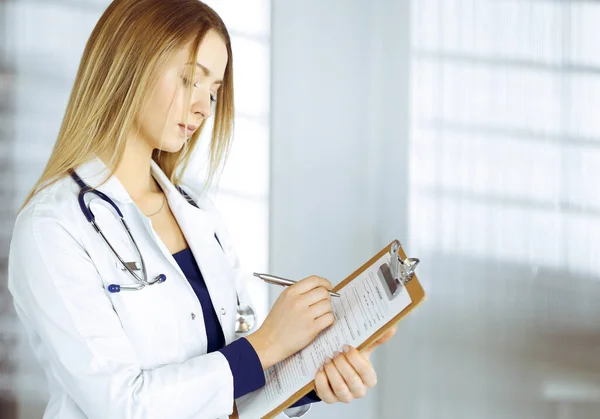 Молодая женщина-врач делает заметки с помощью планшета, стоя в своем кабинете в клинике. Портрет красивой женщины-врача со стетоскопом. Перфектное медицинское обслуживание в больнице — стоковое фото