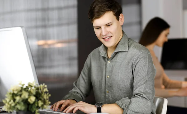 Hard werkende jonge zakenman in een groen shirt is op zoek naar wat informatie op zijn computer, terwijl hij achter het bureau zit in een modern kabinet met een vrouwelijke collega op de achtergrond. Hoofdschot of — Stockfoto