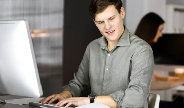 Jeune homme d'affaires et programmeur en chemise verte vérifie un plan d'affaires, tout en étant assis sur le bureau dans un cabinet moderne avec sa collègue féminine sur le fond. Concept de succès — Photo