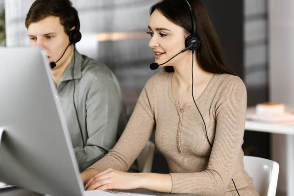 Two young people in headsets are talking to the clients, while sitting at the desk in an office. Focus on woman. Call center operators at work