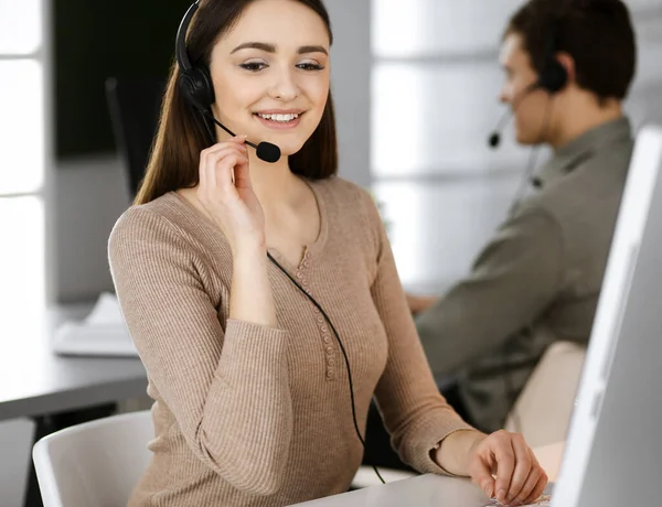 Fiatal barátságos lány fülhallgatóban beszél egy cég ügyfelével, miközben egy modern irodában ül a kollégájával. A munkahelyi call center üzemeltetői — Stock Fotó