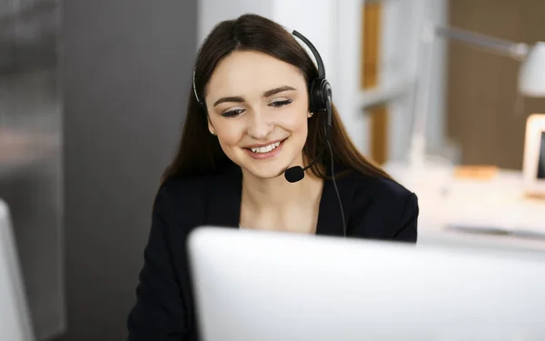 Jeune fille amicale dans les écouteurs parle à un client des entreprises, tout en étant assis sur le bureau dans un bureau moderne avec son collègue. Opérateurs de centre d'appels au travail — Photo