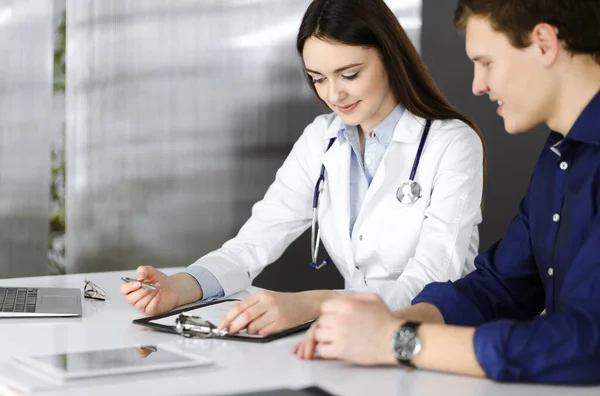 Amichevole giovane donna-medico sta ascoltando la sua paziente, mentre sono seduti insieme alla scrivania in un armadio. Il medico sta tenendo in mano una cartellina per aver riempito i farmaci. — Foto Stock