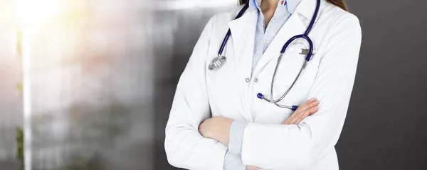 Okänd ung kvinna läkare står med armarna korsade i solig klinik. Medicinskt koncept under Coronavirus pandemi. Mynt-2019 — Stockfoto