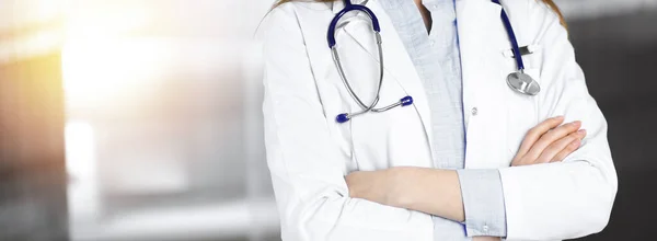 잘 알려 지지 않은 젊은 여성 의사가 양지바른 병원에서 팔짱을 끼고 서 있습니다. 코로나 바이러스 대유행 동안의 의학적 개념이다. Covid-2019 — 스톡 사진