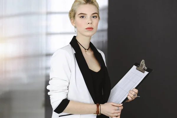Young blonde business woman or female student in white suit is standing in modern office. Lifestyle and diverse people concept