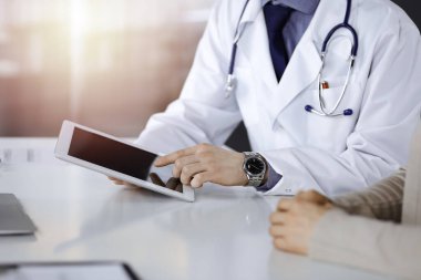 Bilinmeyen bir erkek doktor ve hasta kadın karanlık bir klinikte tablet bilgisayar kullanırken bir şeyi tartışıyor, arka planda ışık parlıyor. Hastanedeki en iyi sağlık hizmeti, ilaç, salgın durdurma.