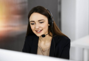 Kulaklık takmış genç bir kız, güneşli bir ofiste masada otururken bir firma müşterisiyle konuşuyor. İş yerindeki çağrı merkezi operatörleri